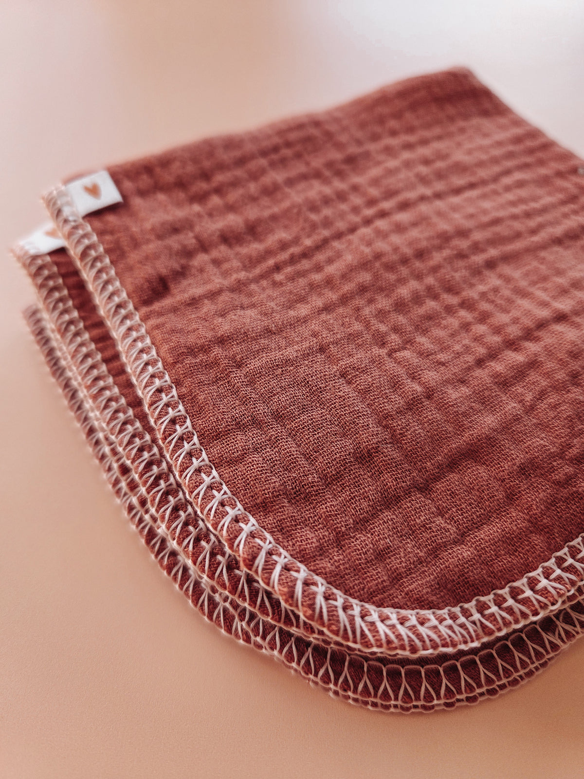 A stack of textured muslin baby wipes in a dusty rose-brown color with white stitched borders, shown on a soft peach-beige background.