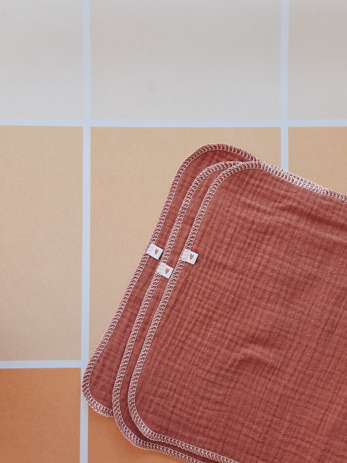 A close-up of a clay colored muslin cloth with a white serged stitched border, laid diagonally on a tiled surface of beige and orange square tiles.