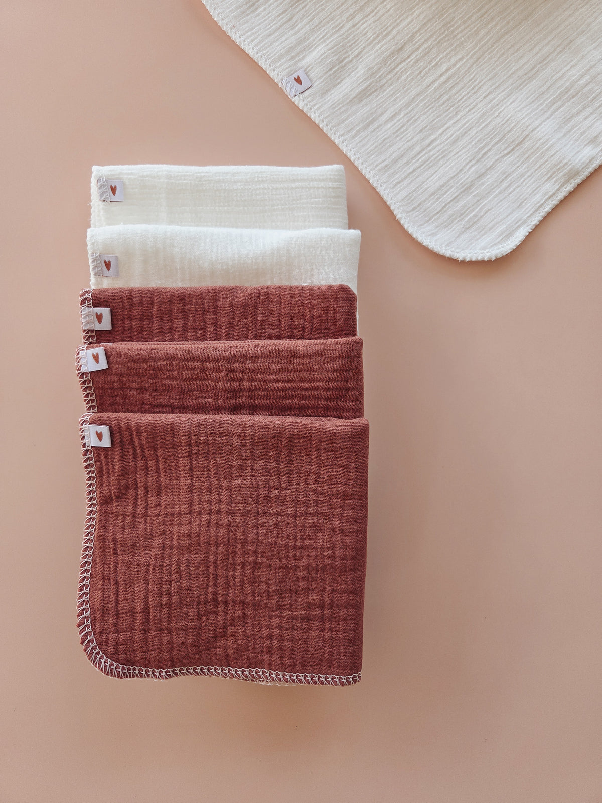 five folded muslin baby wipes in cream, clay, and rust shades, arranged vertically on a warm peach background with a partial sixth wipe visible in the top right corner.