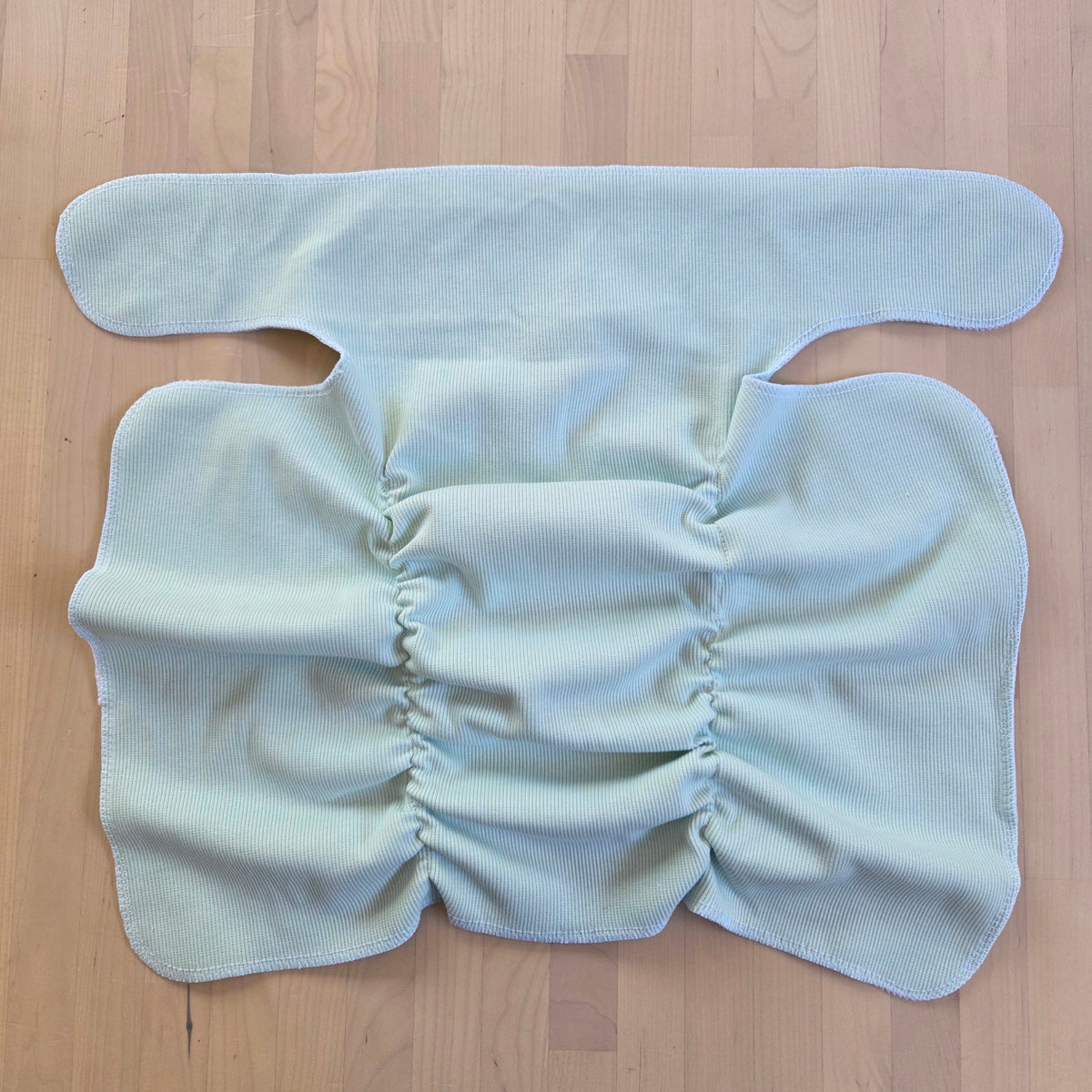 A light mint reusable cloth diaper laid flat on a light wood floor, showing its elasticized features